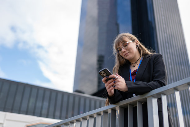 Woman texting outdoors leaning on a railing with modern city skyscrapers in the background图片下载