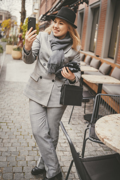穿着灰色西装、围巾、帽子和手套的商界女性，在秋季城市的户外咖啡馆露台上行走，一手拿着智能手机，脸上带着笑容。生活方式、商业街、通讯概念。图片下载
