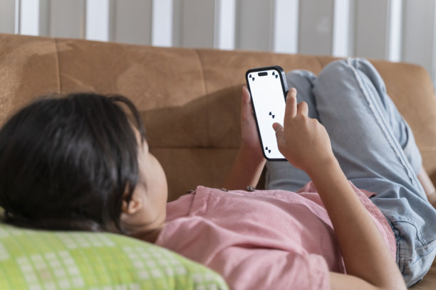 专注于使用智能手机的亚洲十几岁女孩，躺在棕色沙发上，配有用于科技和社交媒体的样机屏幕图片下载