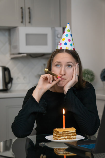 一个戴着派对帽的悲伤孤独的女人，正对着笔记本电脑吹响派对喇叭庆祝生日，她坐在厨房桌旁，桌上放着生日蛋糕和点燃的蜡烛。竖式照片图片下载