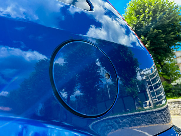 这张特写照片展示了一辆蓝色汽车，映照出城市天际线。其油箱盖突显了现代设计，将鲜艳的色彩与云彩和绿植融为一体，象征着都市生活和旅行。图片下载