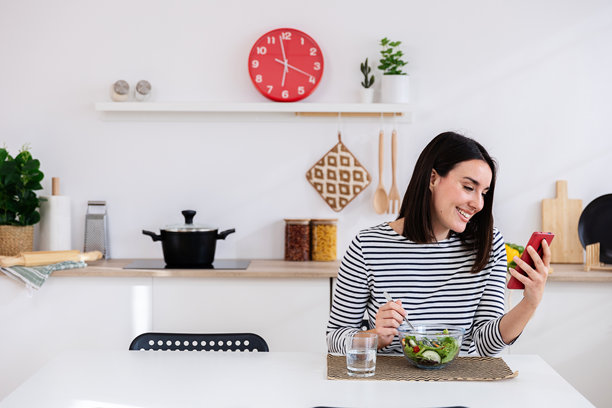 Smiling woman eating fresh salad and browsing social media on phone in kitchen图片下载