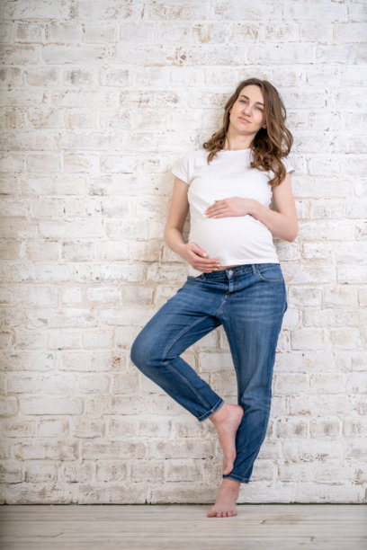 身穿休闲牛仔裤和白色T恤的孕妇在白色砖墙前摆姿势图片下载