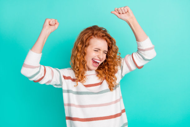 一位留着姜黄色波浪卷发的年轻女子，双手举起，穿着毛衣，在青色的背景前欢呼的照片图片下载