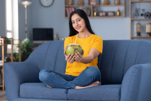 穿着黄色T恤的可爱小女孩在家里的沙发上喝椰子水，照片素材图片下载