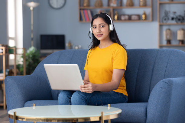 照片：可爱的年轻女孩戴着耳机在家里的沙发上，身穿黄色T恤，进行远程视频通话图片下载