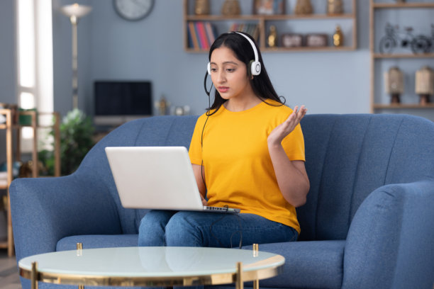 照片：戴着耳机的可爱小女孩身穿黄色T恤，在家中的沙发上进行视频通话的素材照片图片下载