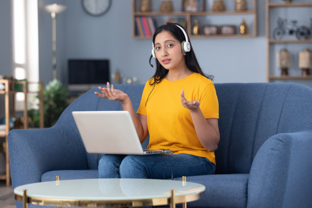 照片：一名戴着耳机、穿着黄色T恤的可爱小女孩在家里的沙发上进行视频通话的素材图片图片下载