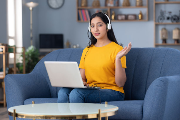 一名戴着耳机的可爱小女孩，身穿黄色T恤，在家里的沙发上进行视频通话的素材照片图片下载