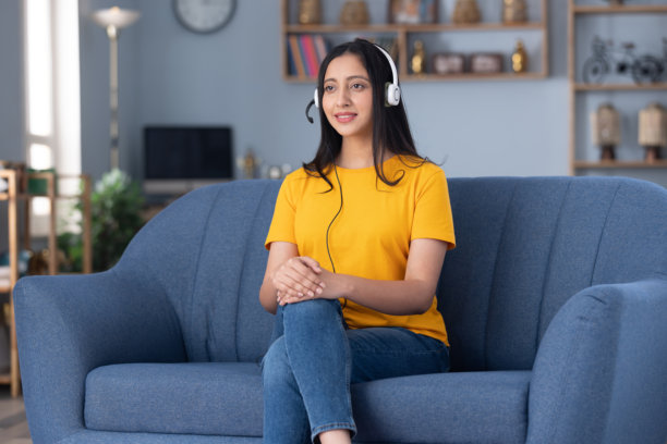 照片：可爱年轻女教师戴着耳机，穿着黄色T恤，在家里的沙发上坐着图片下载