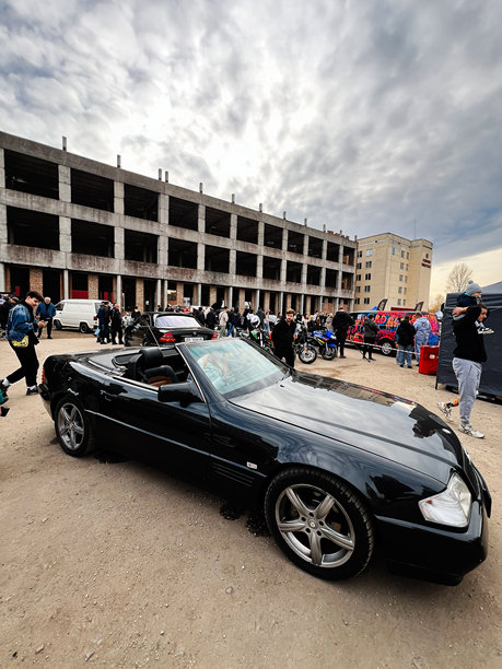 一辆黑色的梅赛德斯-奔驰SL级R129敞篷跑车，在户外汽车展上，背景是人群和未完工的建筑，工业场地图片下载