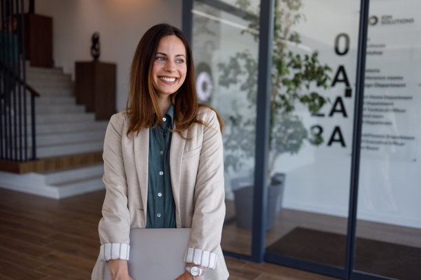 自信的女商人在现代办公楼大堂站立，手持笔记本电脑，面带微笑图片下载