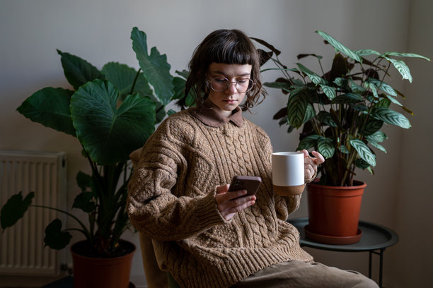 一个少女坐在椅子上，手中拿着茶杯，一边看新闻，一边浏览照片，一边用智能手机发消息图片下载