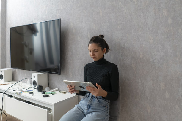 一位女士坐在地板上，在客厅里专注地看着平板电脑，旁边还有一台电视。图片下载