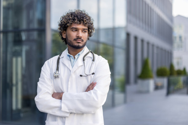 一名年轻的印度男性医生近距离照片，身穿白色制服，戴着听诊器，双臂交叉站立，看向一旁图片下载