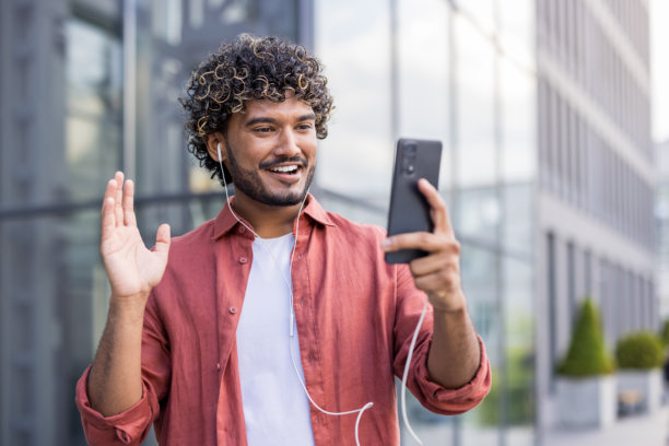 一名戴着耳机的年轻印度男子在户外，通过手机进行视频通话，并向镜头打招呼的特写照片图片下载