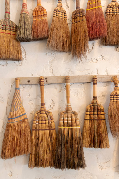 手工编织草扫帚，手柄处有编织纹理，挂在乡村风格的墙壁上，色调温暖，散发着复古手工魅力。图片下载