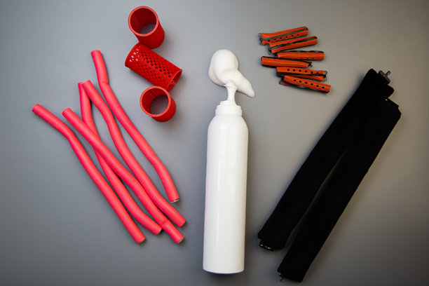 在灰色背景上的特写镜头展示了一瓶用于长期定型的美发摩丝，其中倾倒出白色泡沫，旁边有各种形状和尺寸的卷发器。造型、发型、卷发的概念。图片下载