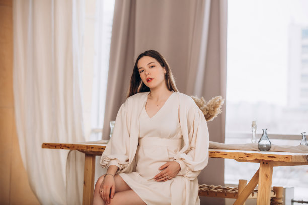 一位留着长发、身穿优雅米色连衣裙的年轻美丽孕妇模特在家中图片下载