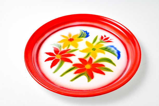 一个带有鲜红色边缘的圆形复古搪瓷盘子或托盘。中心以白色为背景，饰有色彩鲜艳的花卉图案，包括红色和黄色的花朵，以及绿色和蓝色的点缀。图片下载