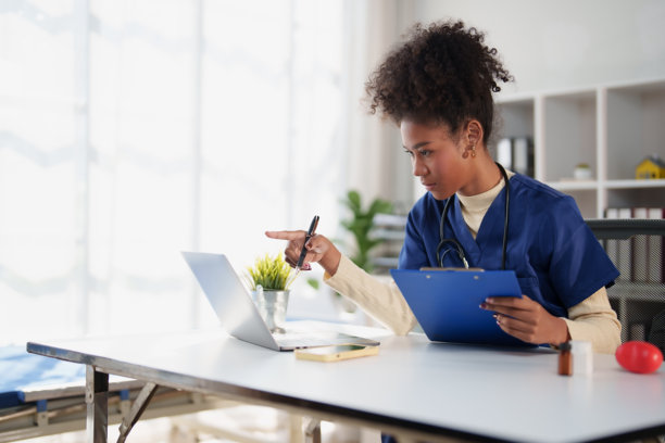 一位非裔美国医疗专业人士正在现代医疗办公室的办公桌前，指着笔记本电脑屏幕，并拿着一个文件板，准确地查看患者数据和记录。图片下载