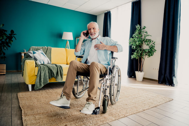 一位坐着轮椅的老年男子在明亮的客厅里用黄色的沙发、绿植和舒适的装饰品打电话，突出了家庭生活中的包容性和多样性。图片下载