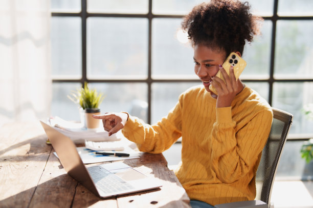 年轻女子在家远程办公，一边用智能手机打电话，一边用手指着笔记本电脑屏幕，强调沟通与效率图片下载