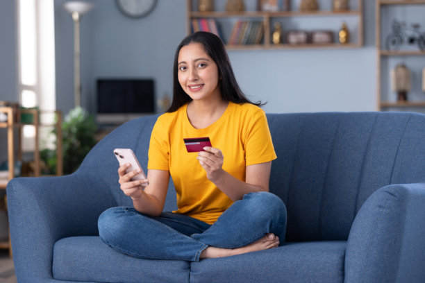 年轻漂亮的女孩在家中沙发上穿着现代服装，一边喝咖啡一边在线购物的库存照片图片下载