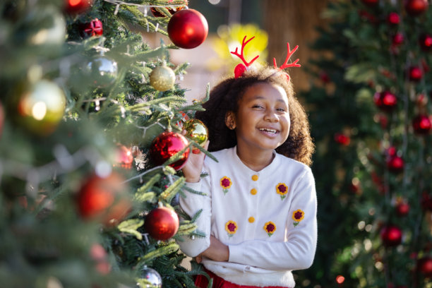 一个快乐的小女孩戴着驯鹿角，在装饰好的圣诞树旁兴奋地微笑。这张图片反映了童年的快乐、节日气氛、假日幸福以及圣诞庆祝的魔力。图片下载