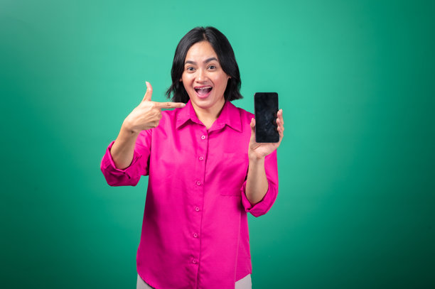 一位穿着亮粉色衬衫的亚洲女性，脸上带着灿烂的笑容，兴奋地举着一部智能手机，指向屏幕，背景为绿色。图片下载