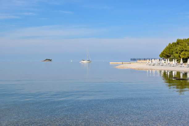 这张照片展示了罗维尼的孤湾，海水平静清澈，一艘帆船停泊在近海，宁静的鹅卵石海滩上摆放着躺椅，天空湛蓝明亮。图片下载