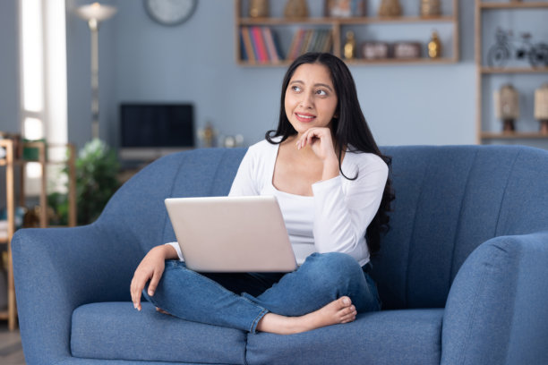 照片：身穿现代服装的漂亮年轻女孩在家中坐在沙发上使用笔记本电脑图片下载