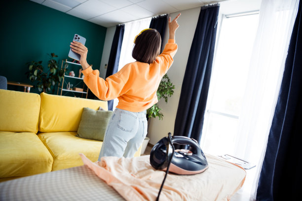 一位穿着橙色毛衣的年轻女子，在阳光明媚的日子里，于明亮的客厅中享受居家休闲时光，并在现代公寓里用沙发作为背景自拍。图片下载