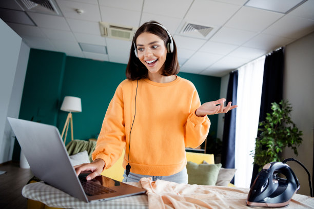 年轻女子戴着耳机，在明亮的客厅里用笔记本电脑在家工作，营造出一种休闲而时尚的居家办公氛围。图片下载