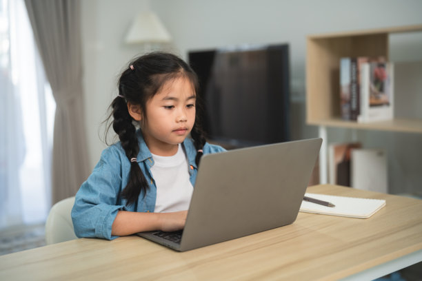 身处舒适的居家环境，身穿休闲蓝色牛仔衬衫的年轻女孩正专注地盯着笔记本电脑屏幕，坐在木制书桌前学习。图片下载