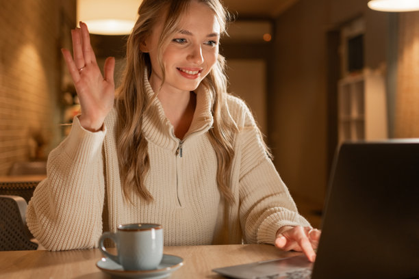 年轻女子穿着毛衣，在咖啡馆参加视频通话或虚拟会议，手中端着咖啡，享受着晚间时光，并向屏幕挥手。图片下载