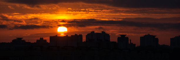 现代都市上空壮丽的红色日落。在多云发光的天空中，太阳圆盘低悬在建筑之上。美丽的夜晚城市景观照片。15x5格式宽屏全景图片下载