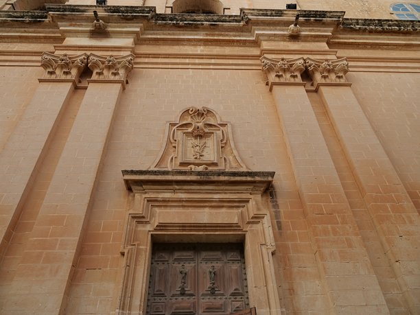 马耳他古老城堡城市姆迪纳圣保罗大教堂的入口外观。图片展示了精美的巴洛克风格石灰石建筑、巨大的柱子以及主入口上方带有纹章的装饰，背景是晴朗的蓝天和几朵白云。图片下载