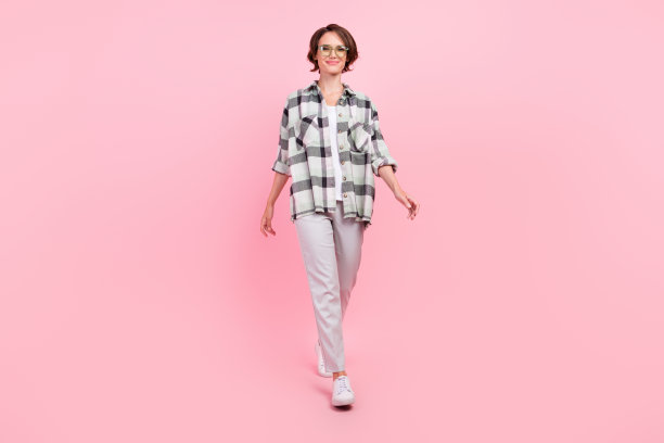 戴着眼镜、衬衫、裤子和运动鞋的漂亮年轻棕发女士的全身侧面照片，在粉色背景上孤立图片下载