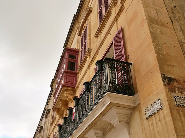 一张垂直拍摄的照片，展示了马耳他古老防御城镇“无声之城”姆迪纳迷人的狭窄街道。图像突出了传统的马耳他石灰岩建筑，其特色是华丽的锻铁阳台和独特的红色封闭木制阳台。图片下载