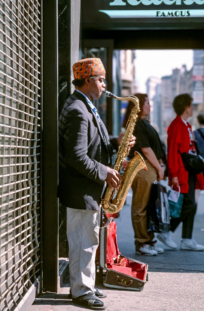 1993年5月9日，美国纽约，一张老式彩色照片，街头音乐家在街边房屋墙壁前演奏萨克斯，路人对此视而不见图片下载