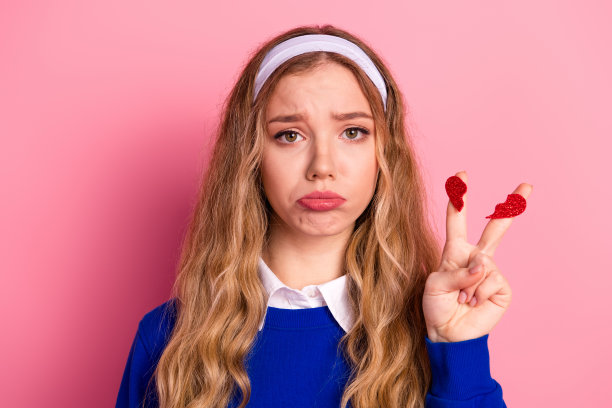 留着金色头发的年轻女孩戴着发带，站在粉色背景前，手持闪光心形，做出胜利手势，用于时尚风格和生活方式推广。图片下载