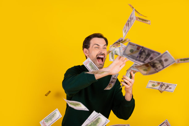 一名穿着毛衣的年轻男子对着背景为明亮的黄色，钱币如雨般落下，正在大笑的素材照片图片下载