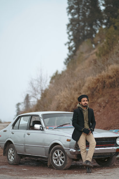 一位留着胡须的男子，身着传统帽子和黑色外套，在冬天崎岖的山路上倚靠在一辆银色汽车旁。图片下载