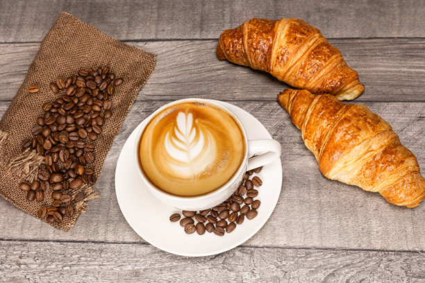 木质背景上的卡布奇诺咖啡，带有花朵拉花、羊角面包和咖啡豆。图片下载