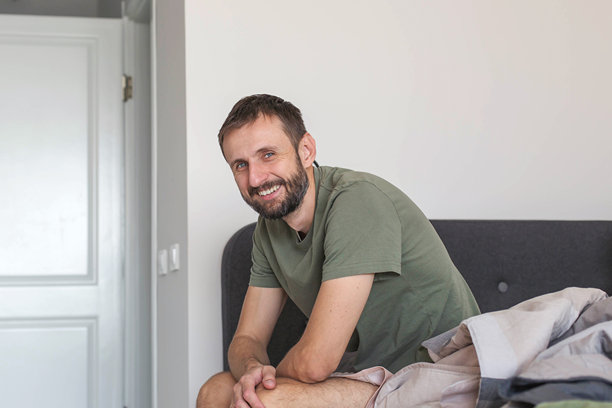 在家床上坐着的成年男子，对着镜头微笑，舒适卧室内的放松休闲生活方式，晨间日常和居家生活概念，自然光线和真实的居家氛围图片下载