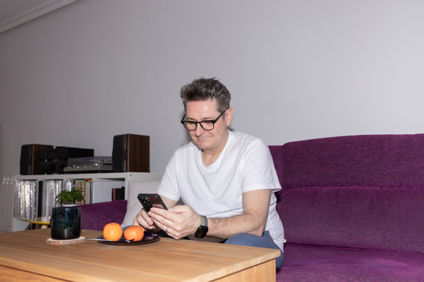 一位50多岁的男子在家里的客厅沙发上放松，旁边桌子上放着咖啡和橘子，他正在使用智能手机，这是真实生活场景下的居家休闲和日常生活。概念是数字生活和日常居家生活。图片下载