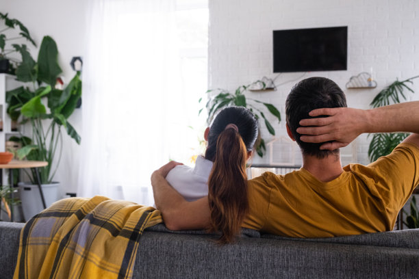 一对情侣在家中白色内饰的沙发上观看电视，电视屏幕为黑色，模拟场景。居家休闲、放松、爱、家庭、约会。图片下载