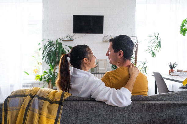 一对夫妇在家中白色内饰的沙发上观看电视，电视屏幕为黑色模拟效果。居家休闲、休闲娱乐、爱情、家庭、约会图片下载