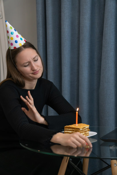 封锁期间，一位戴着派对帽的年轻女子在网上庆祝生日，她看着笔记本电脑，手里拿着一块点燃生日蜡烛的生日蛋糕。竖幅照片。图片下载